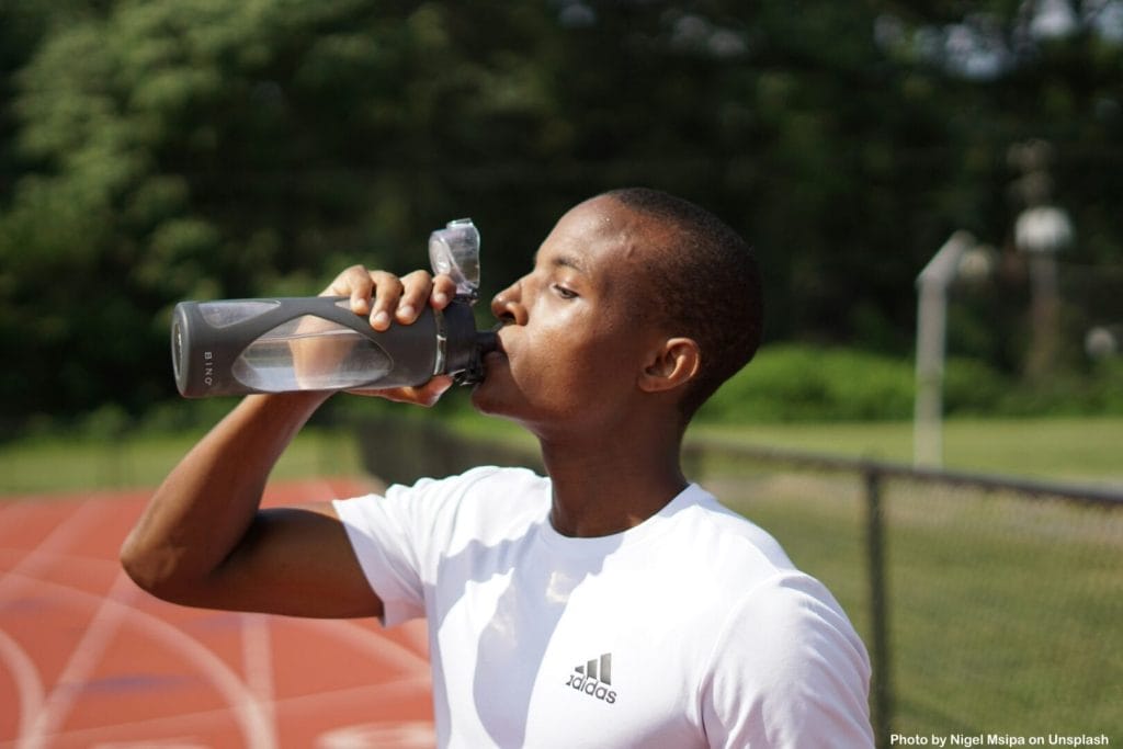 A Black person with very short hair wearing a tight white t-shirt. They are drinking water from a glass water bottle that they hold it in their right hand. A track is in the background.