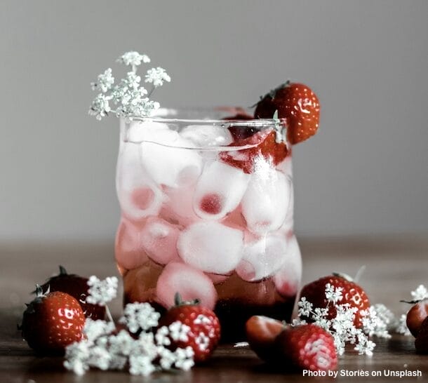 Drink more water with this strawberry iced water. The glass is full with oval ice cubes and several strawberries are in and around the glass. Small white flowers are mixed in with the berries.