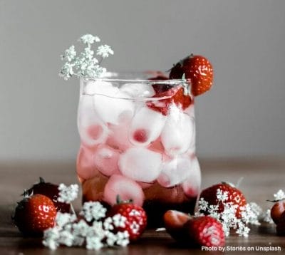 Drink more water with this strawberry iced water. The glass is full with oval ice cubes and several strawberries are in and around the glass. Small white flowers are mixed in with the berries.